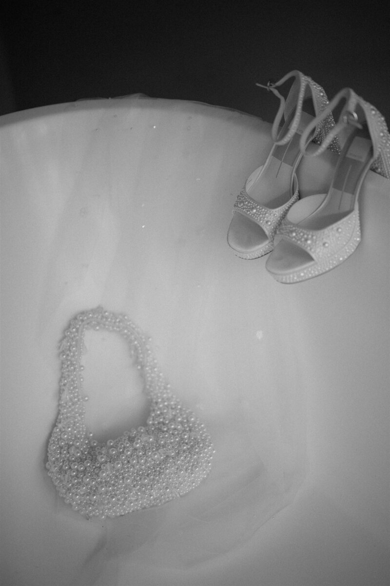 Wedding shoes and a beaded bag resting on a wedding dress, in black and white